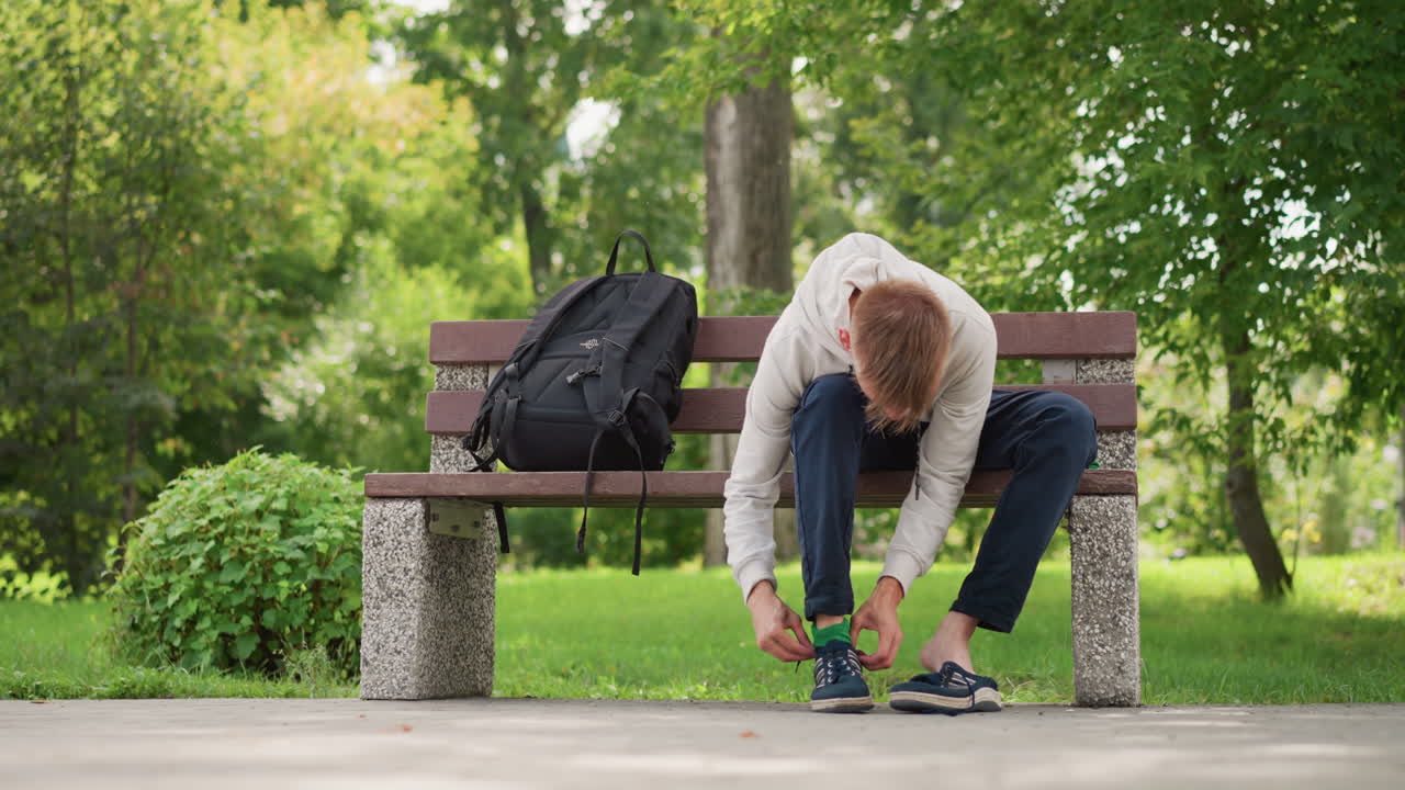 햇볕이 잘 드는 공원의 벤치 옆에서 백팩을 메고 신발 끈을 묶는 백인 남성. 젊은 백인 남성이 끈을 묶기 위해 몸을 숙여 끈을 묶고 일어서서 무게를 실어 핏을 확인하며 준비하는 모습이 연상돼요.