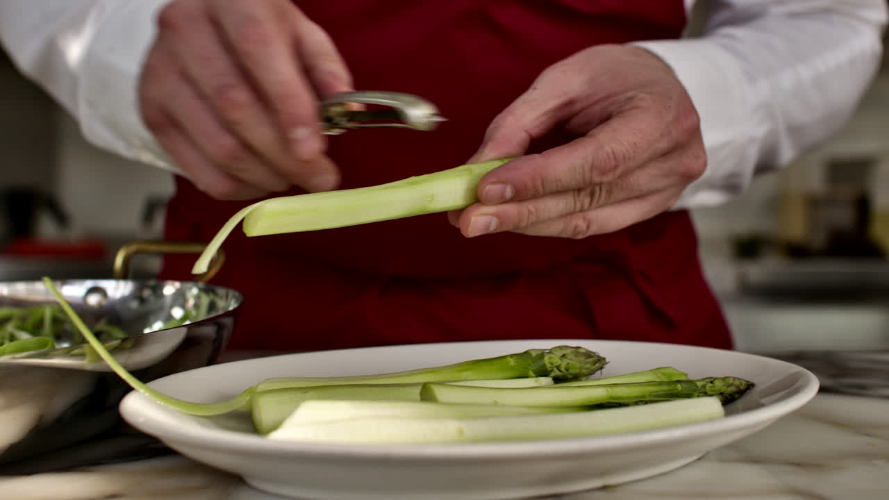 Close-up shot of a chef's hands using a peeler on a fresh green asparagus over a white plate. Culinary prep for a fresh vegetable salad in a generic kitchen setting