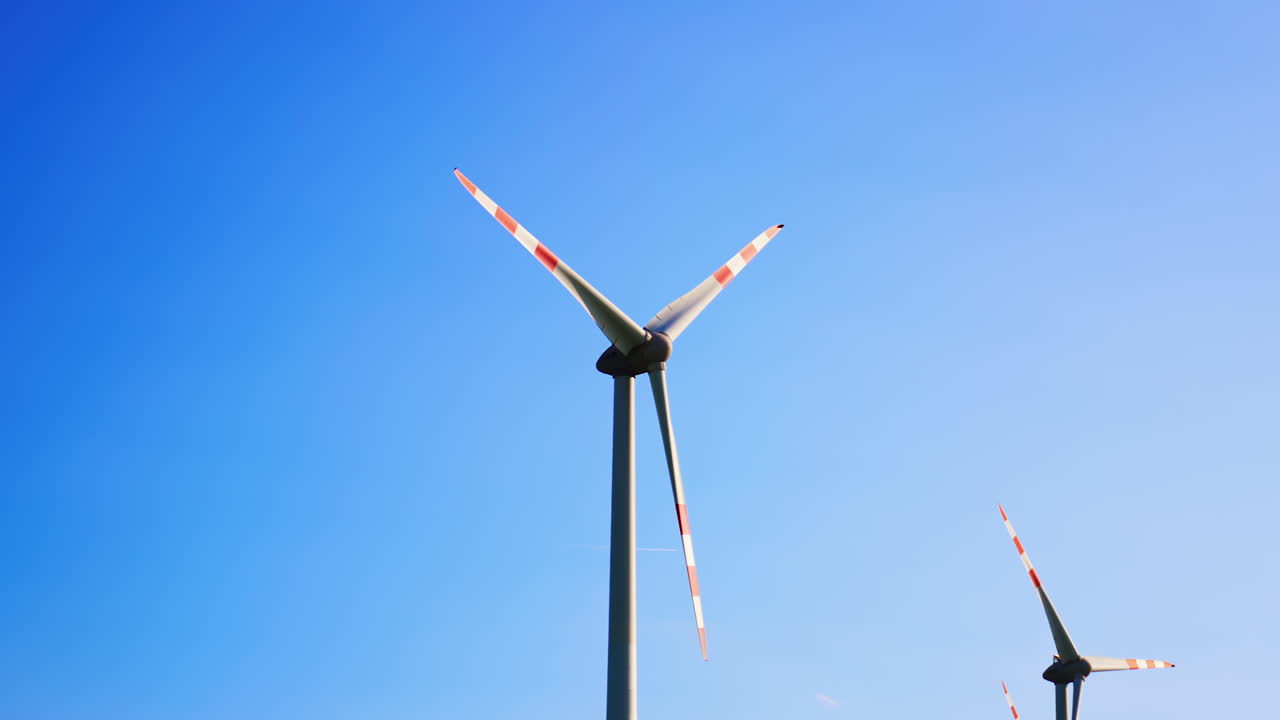 Wind turbines in blue skies. Two wind turbines with red and white blades stand tall against a bright blue sky, showcasing renewable energy in action