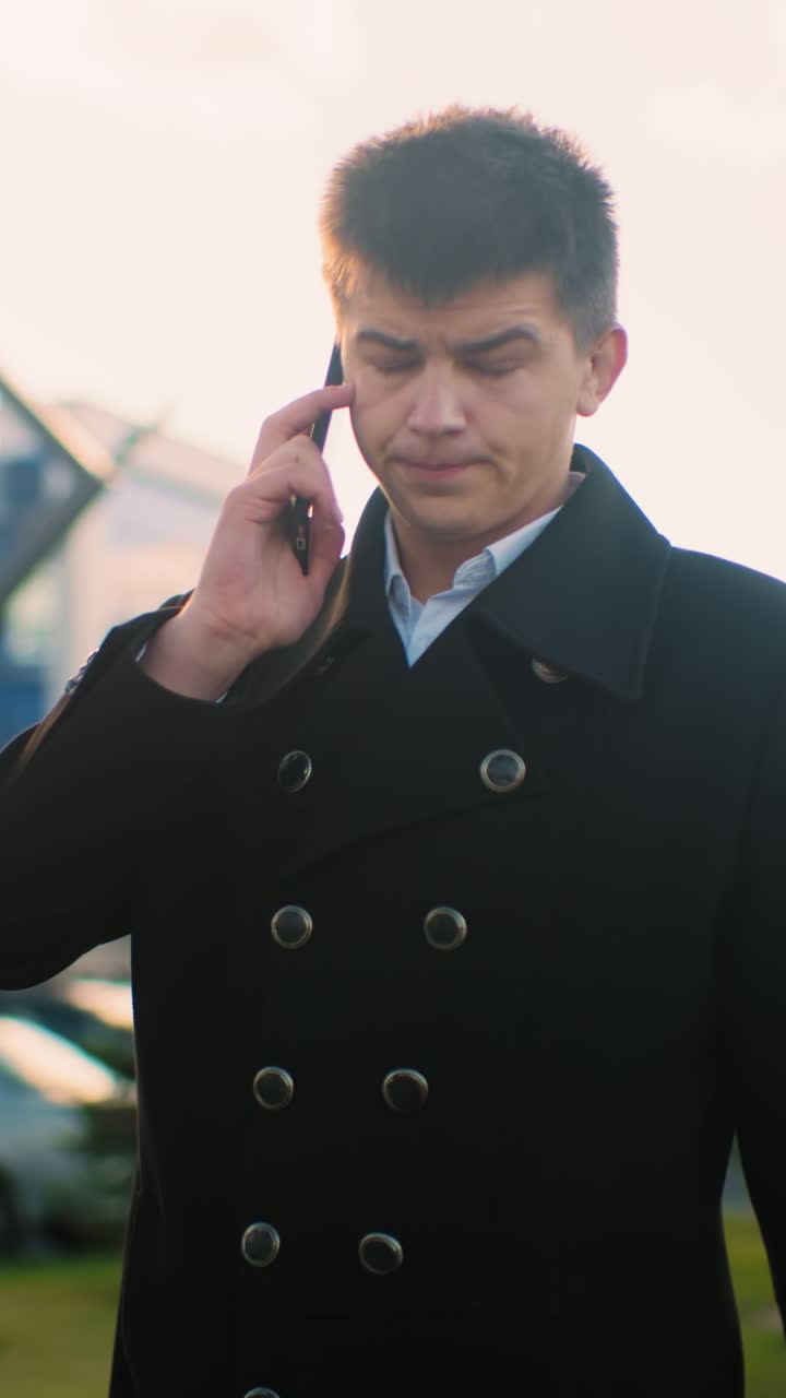 검은 코트를 입은 사업가가 전화 중에  에 전화를 들고, 현대적인 건물, 주차된 자동차, 그리고 배경의 초록색으로 도시 지역에서 밝은 빛 아래 자신감 있게 고 있습니다.