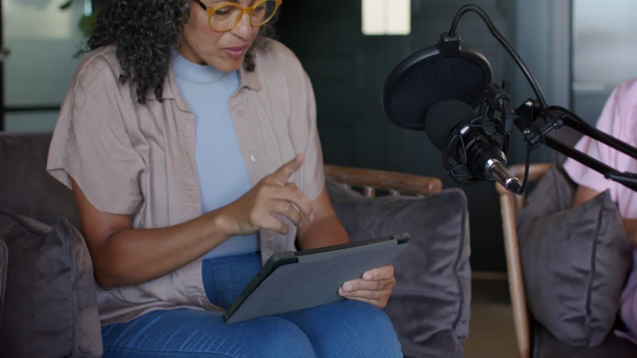 Due podcaster donne su un divano in studio che regolano il microfono dopo lo spostamento di un filtro anti-pop, mentre toccano un tablet.