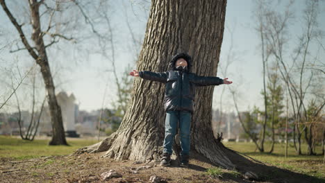 un niño joven está de pie con la espalda contra un gran tronco de árbol, mirando hacia arriba con los brazos extendidos ampliamente, está vestido con una chaqueta negra brillante, vaqueros y botas, con árboles a su alrededor