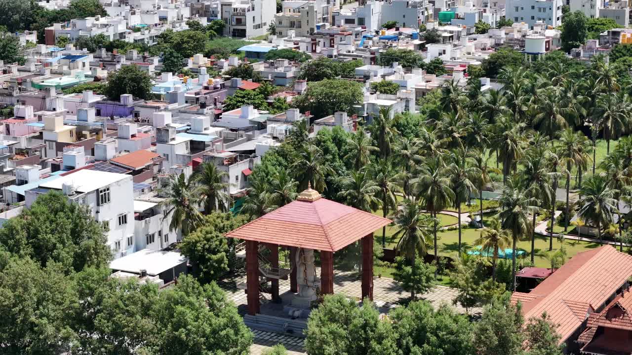 Aerial view of a white statue of a seated Anjaneya Temple sheltered under a traditional pavilion (mandapa) with a tiled roof. The statue sits in a large, paved courtyard with green coconut trees