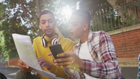 dos felices amigos de raza mixta sentados, usando un teléfono inteligente y un mapa