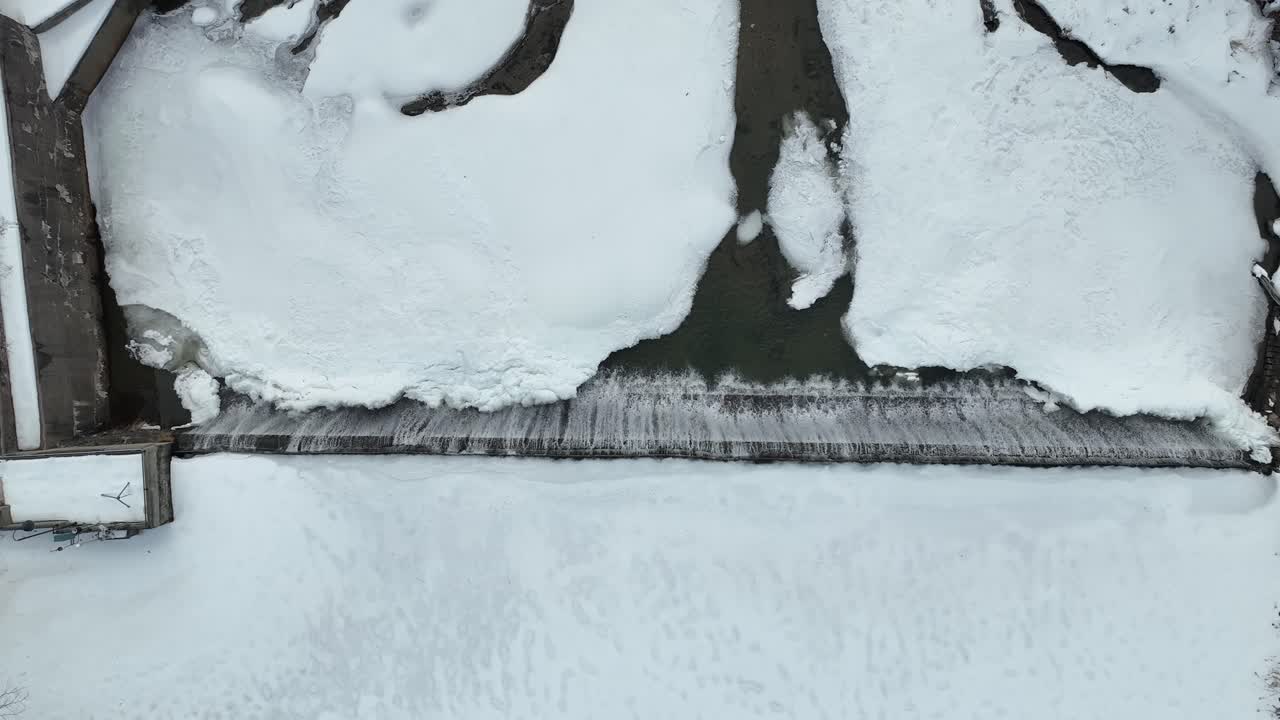 Frozen dam with water flowing over, aerial winter landscape with icy river and snow