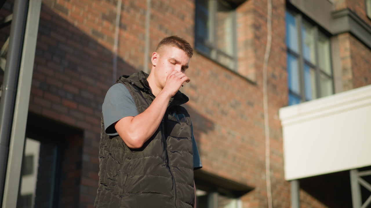 young boy dressed in black vest and blue shirt raises cigarette to lips with intent to smoke then suddenly changes mind and discards it outdoors against brick building