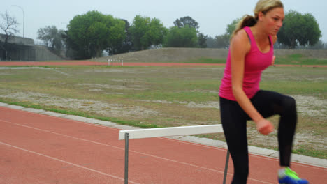 Side-view-of-Caucasian-female-athlete-jumping-over-hurdle-on-race-track-at-sports-venue-4k