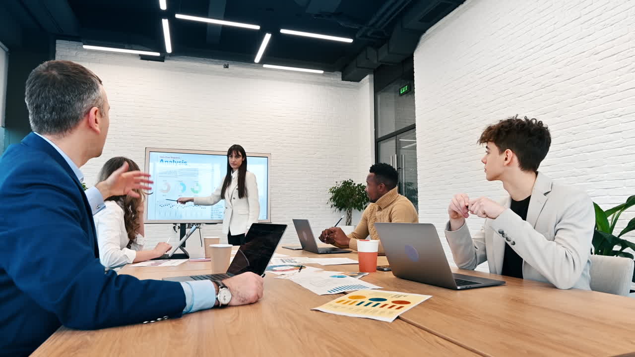 Líder de equipe feminina em reunião de negócios em um escritório, discutindo assuntos de negócios com funcionários usando um display grande com gráficos, documentos e dispositivos eletrônicos na mesa