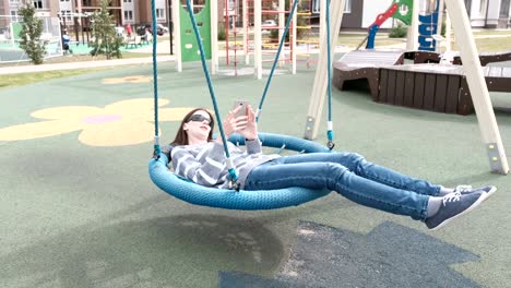 woman is swinging on empty webbed swing and typing a message on mobile phone.