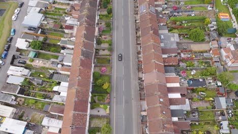 Aerial-view-of-a-black-car-travelling-down-a-terraced-street-in-UK-suburbs,-showcasing-rows-of-identical-houses-with-small-front-gardens-and-backyards