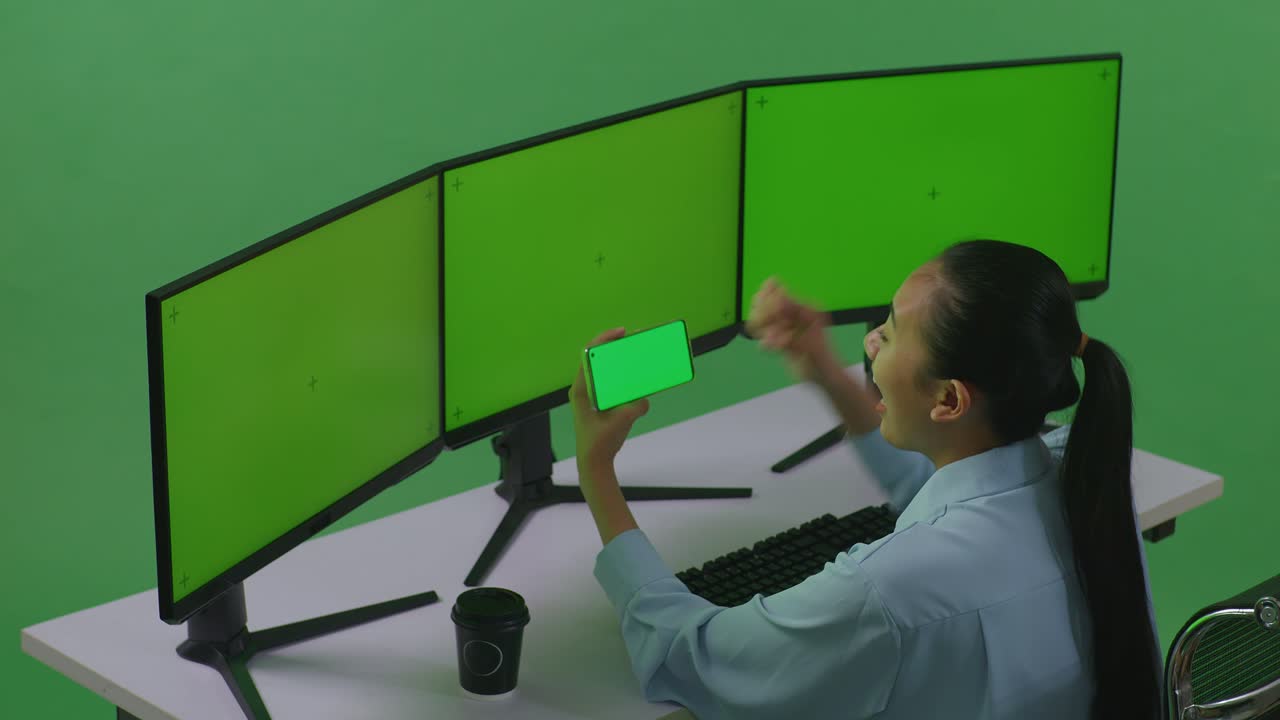 Side View Of Happy Asian Woman Looking At Mobile Phone Green Screen And Celebrating While Working With Multiple Computer Monitor In Green Screen Studio