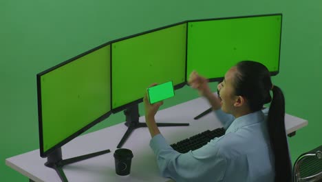 vista lateral de una mujer asiática feliz mirando la pantalla verde del teléfono móvil y celebrando mientras trabaja con múltiples monitores de computadora en el estudio de pantalla verde