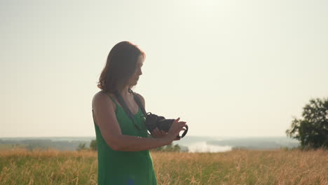 female tourist with eyes closed holds camera ready after removing lens cover while standing in tall golden grass under soft sunlight during quiet summer afternoon in open countryside landscape