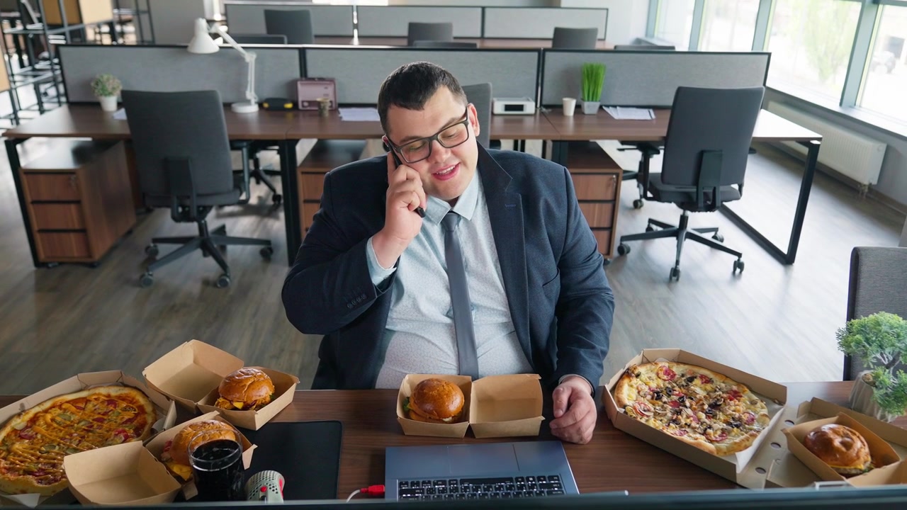 Empresario con sobrepeso trabajando en un portátil y hablando por teléfono con comida rápida
