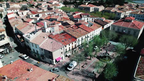 aerial-shot-with-a-drone-over-a-village-in-the-south-of-France-with-red-roofs,-a-small-square-with-a-fountain-and-trees,-cars-driving-by,-birds-flying-by