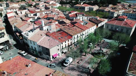 tomada aérea con un dron sobre un pueblo en el sur de francia con techos rojos, una pequeña plaza con una fuente y árboles, coches pasando, pájaros volando