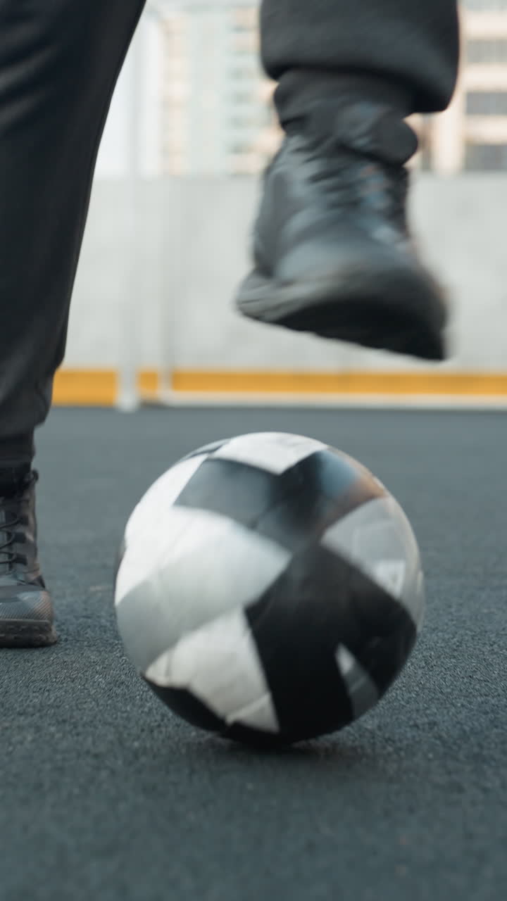 vista de la pierna de un atleta con capucha entrenando hábilmente con el fútbol en la arena deportiva, tocando la pelota hacia adelante con precisión, el fondo incluye una valla en forma de cadena y un edificio residencial alto
