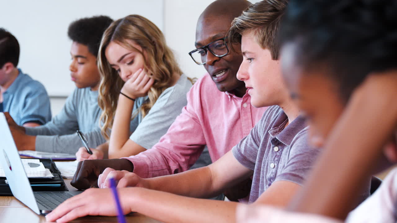 High School Teacher At Desk Working With Pupils Using Digital Devices In Technology Class