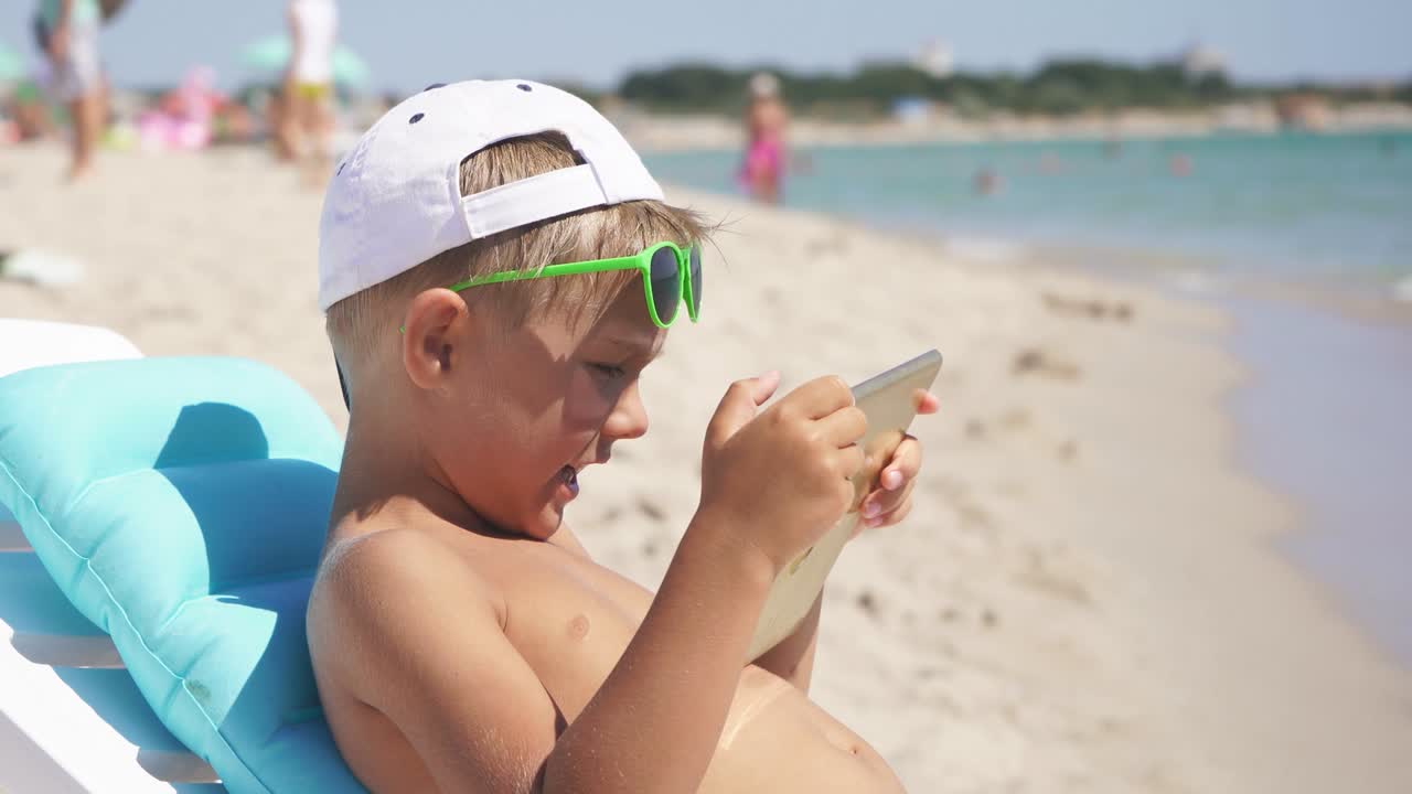 바가에 앉아있는 소년은 빛의 여름날에 태블릿을 사용합니다. 모래 해변에 있는 아이는 바가에서 체이스롱에 앉습니다.
