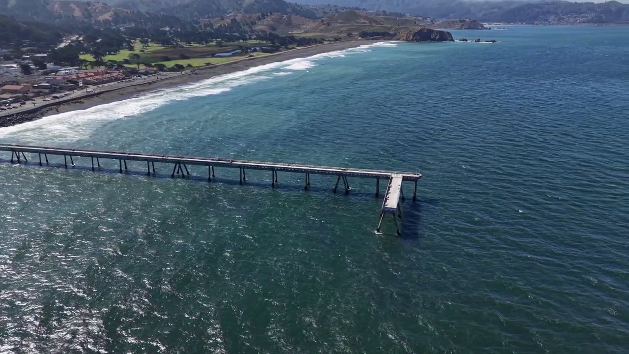 Ampie riprese aeree catturano il molo municipale di Pacifica che si estende in scintillanti onde blu dell'oceano, con la frastagliata costa californiana e colline illuminate dal sole in una drammatica composizione orizzontale.