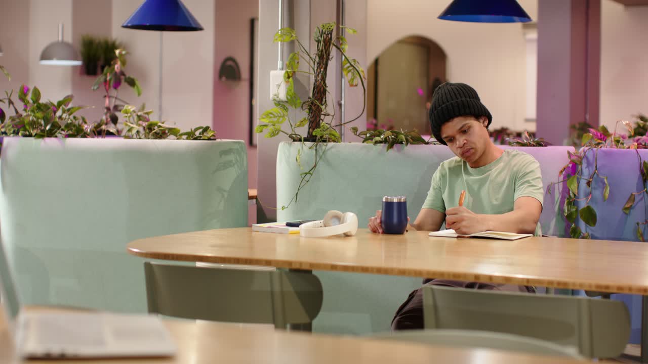 Uomo adulto afroamericano che guarda un taccuino, scrive, tiene una tazza blu in un caffè dai colori pastello, con spazio per il testo.
