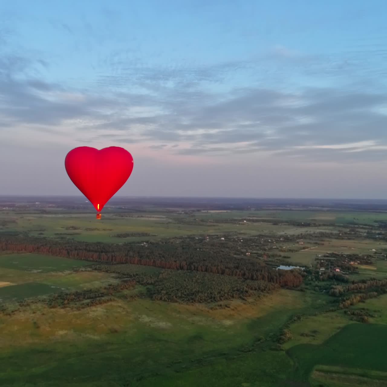 Flight of a red airship. Beautiful hot air balloon in heart shape travelling over the green fields in the morning. Aerostat with basket in the sky.