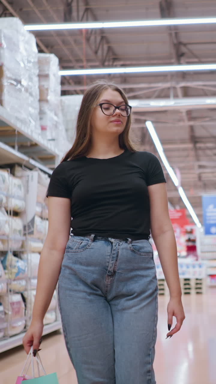 Giovane donna con top nero che cammina vicino allo scaffale di un negozio, osserva i prodotti mentre porta delle buste della spesa, sfondo sfocato con un altro cliente che spinge il carrello