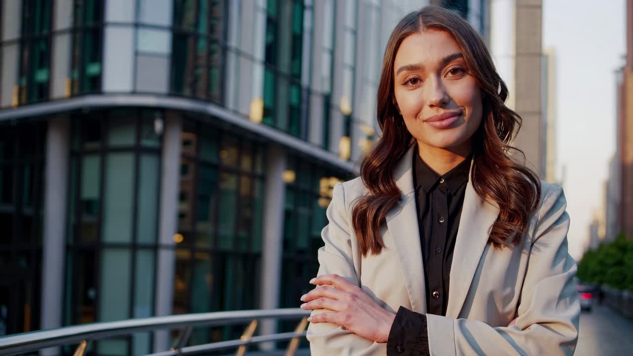 Young businesswoman stands confidently with arms crossed in front of modern office buildings, exuding professionalism and success in the corporate world