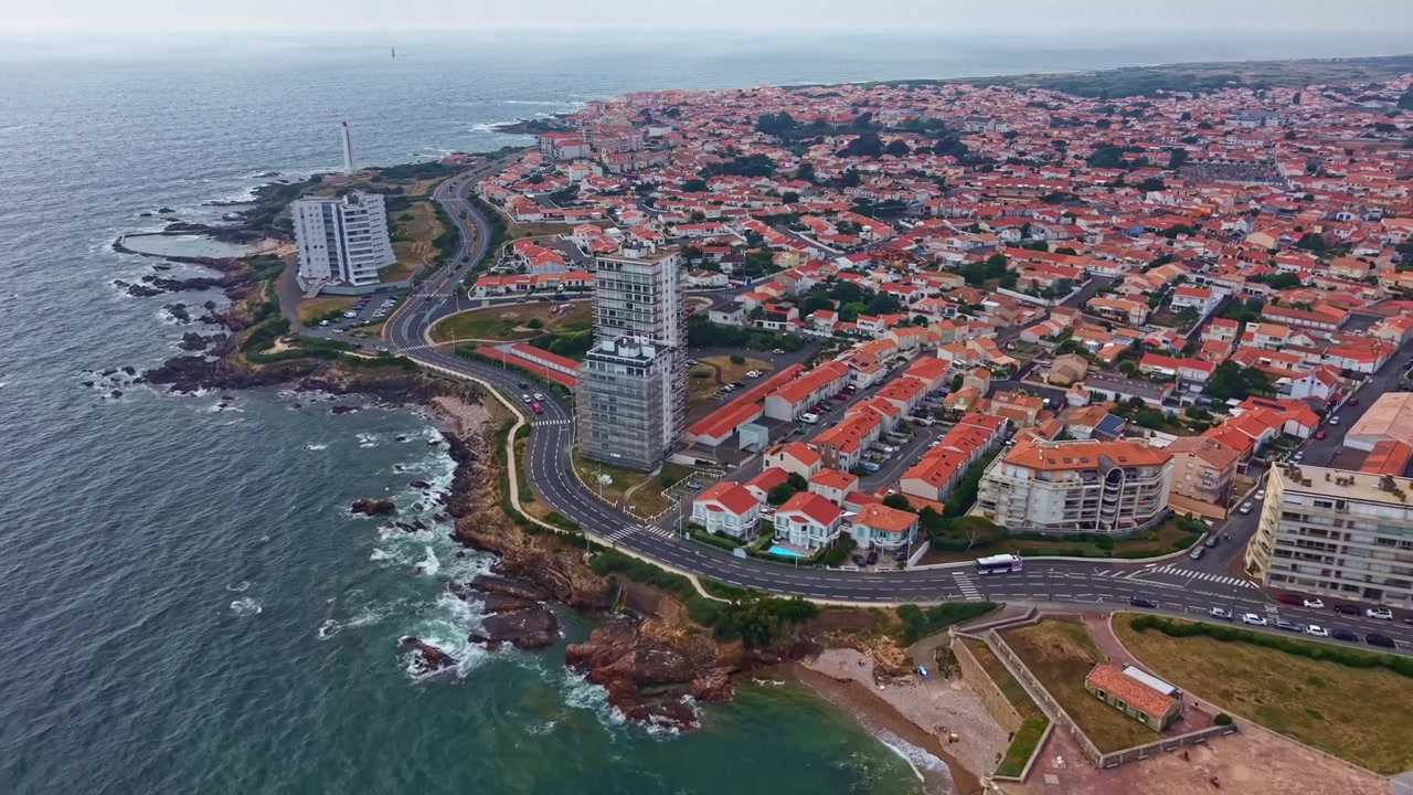 Drone avança pela orla atlântica de Les Sables-d'Olonne, Vendée, revelando o calçadão costeiro, a costa rochosa e o Farol de l'Armandèche ao fundo