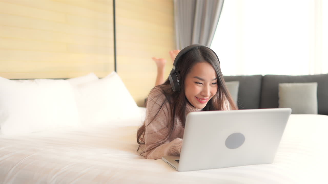 Young Asian woman listening to music and using laptop lying on the bed at hotel room