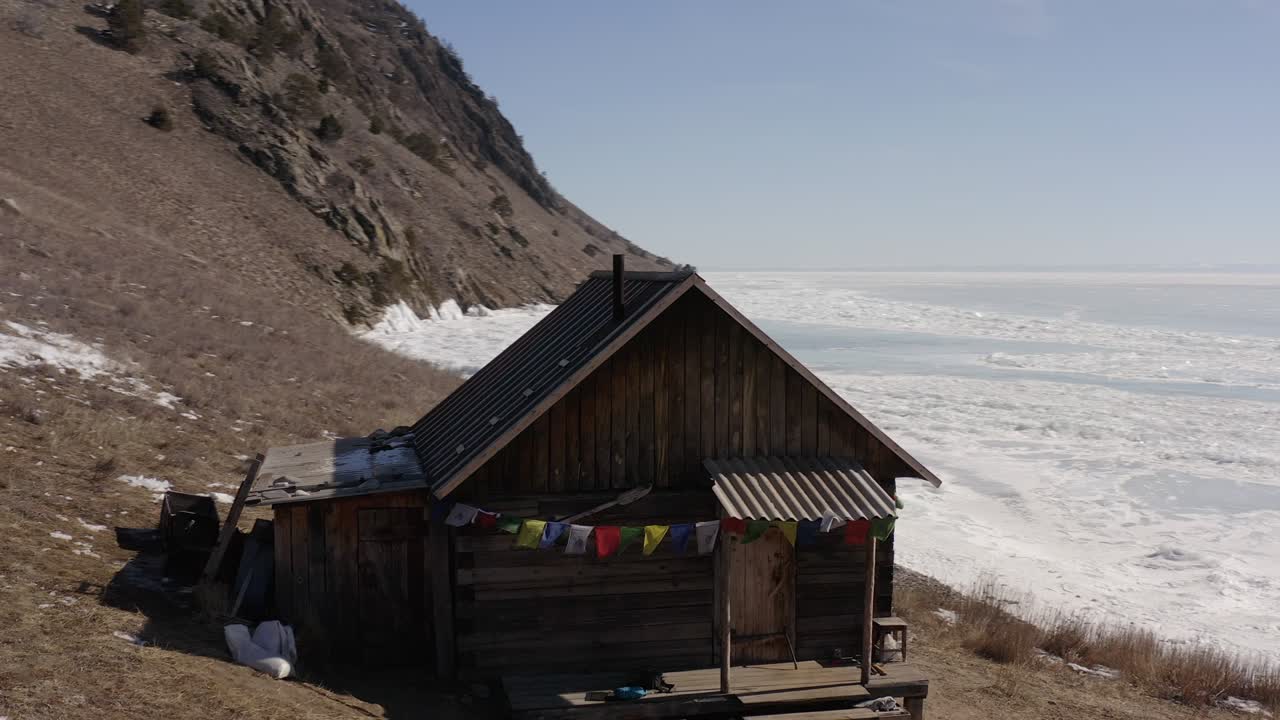 un dron flota sobre una casa con sagradas banderas tibetanas en la orilla rocosa del lago baikal en invierno.