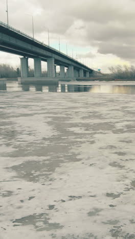 motion over picturesque wide frozen river under grey bridge against cloudy sky on cold winter day aerial view