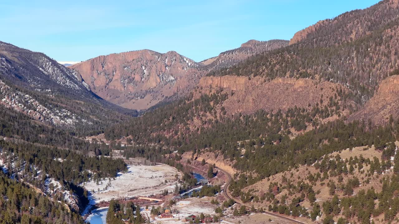 Un volo scorrevole di drone segue un fiume tortuoso in profondità in un aspro canyon di montagna. Fitte foreste sempreverdi coprono i ripidi pendii che conducono a cime alpine rocciose sotto un cielo invernale sereno in 4K