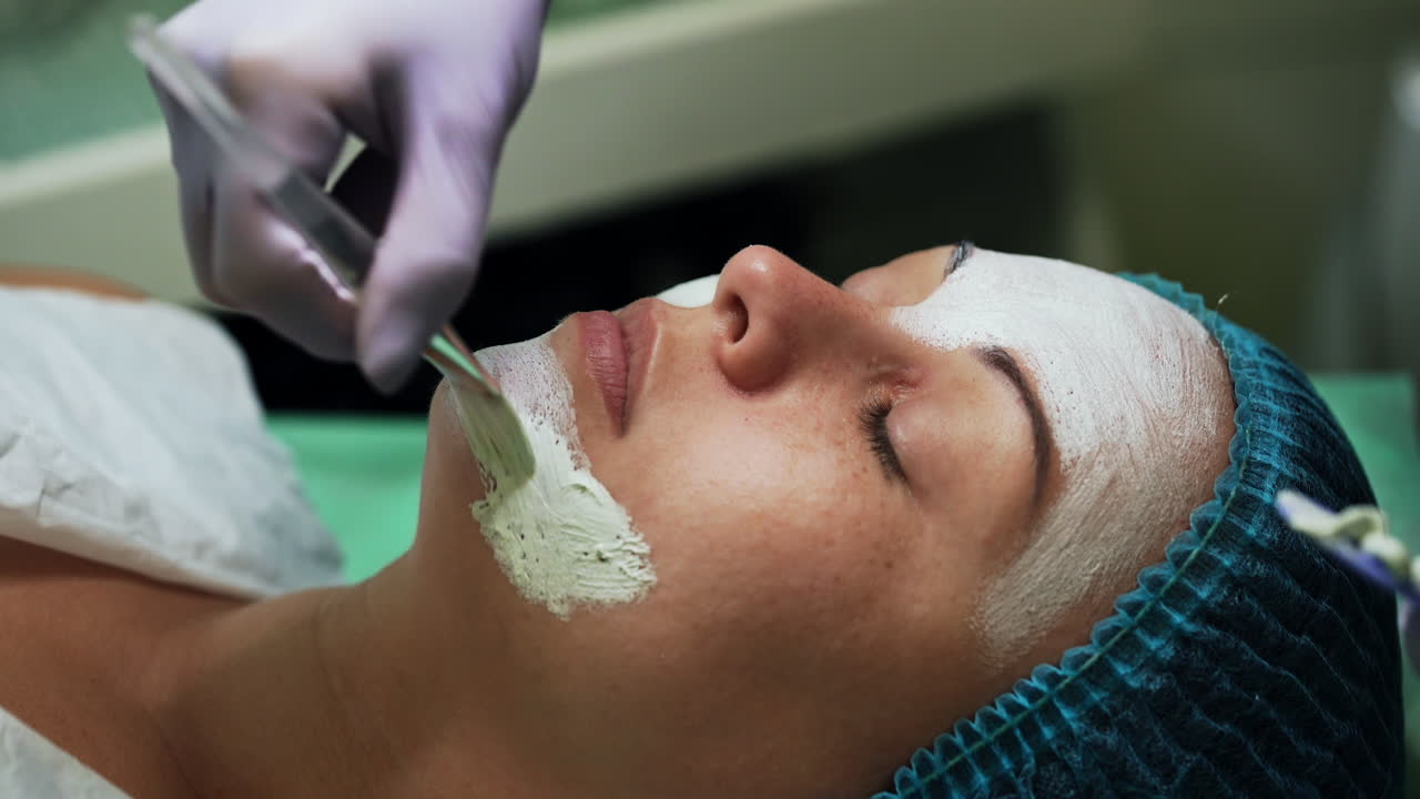 Cosmetologist covering female face with white mask. Beauty specialist uses special brush to apply the mixture. Close up.