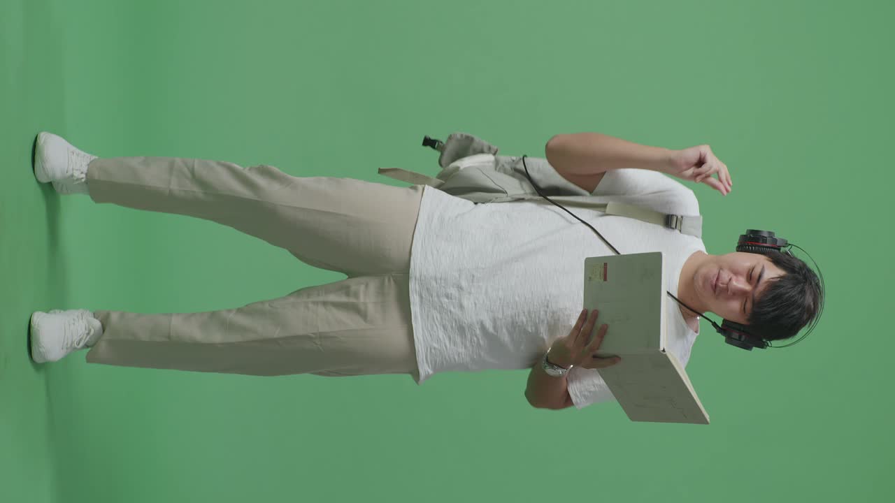 아시아 남성 학생이 책을 들고 헤드폰으로 음악을 듣고 녹색 스크린 스튜디오에서 춤을 추고 있습니다.