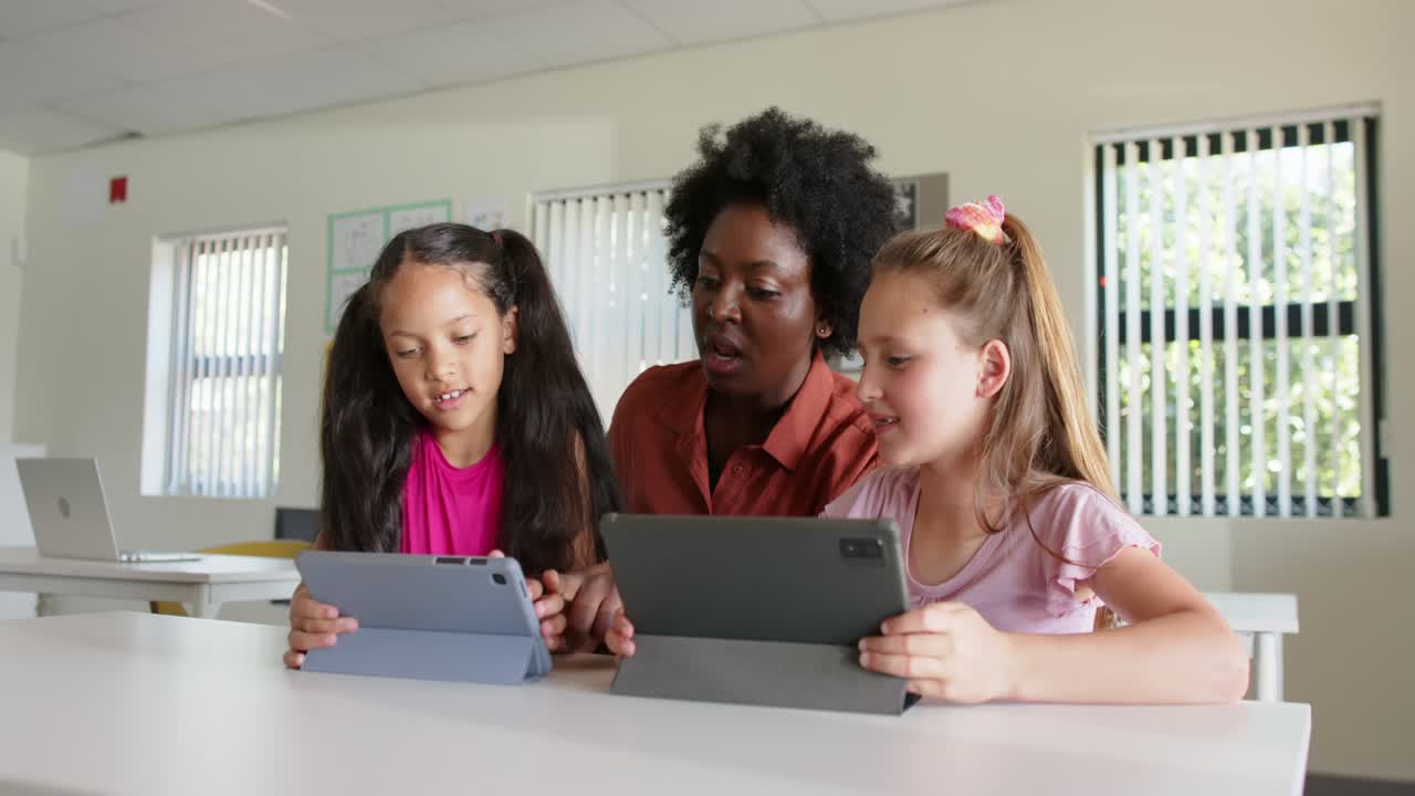 Insegnante donna di diverse etnie che si china, guidando due bambini che usano tablet blu scuri alla scrivania per imparare.