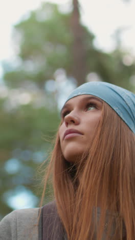 mujer joven caminando en un bosque exuberante, mirando pensativamente hacia abajo, usando bandana azul y sosteniendo correas de mochila, rodeada de vegetación vibrante y árboles altos