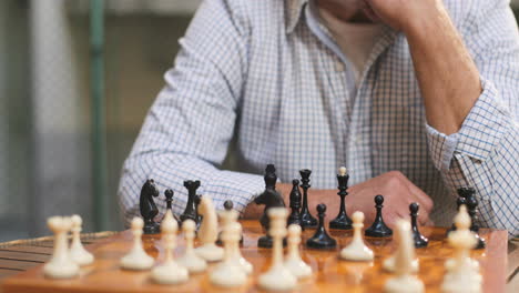 Portrait-Shot-Of-The-Smiled-And-Happy-Good-Looking-Senior-Man-In-A-Cap-And-On-Retirement-Sitting-At-The-Table-With-A-Chess-Board-And-Figures-And-Looking-To-The-Camera