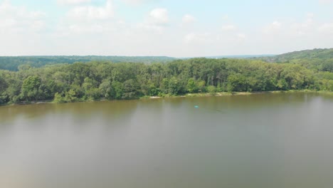 Vuelo-De-Drones-Justo-Sobre-El-Lago-Del-Bosque-En-Illinois-Con-Exuberantes-árboles-Verdes-Y-Cielo-Azul-Nublado-En-Un-Día-Soleado-Mientras-La-Gente-Flota-En-El-Agua-En-Un-Kayak-4k