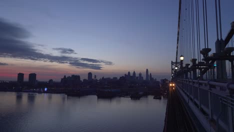 Cruce-De-Tren-De-Metro-Que-Viaja-En-El-Puente-Ben-Franklin-Sobre-El-Río-Delaware-Con-Edificios-Frente-Al-Mar-En-El-Fondo-Al-Atardecer---Plano-General