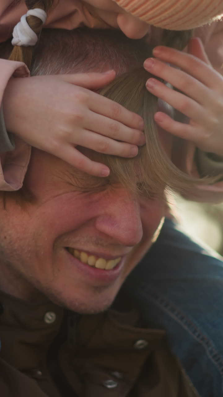 Father with a warm smile as his little girl in a pink jacket playfully places both her hands and head on his, outdoors at sunset