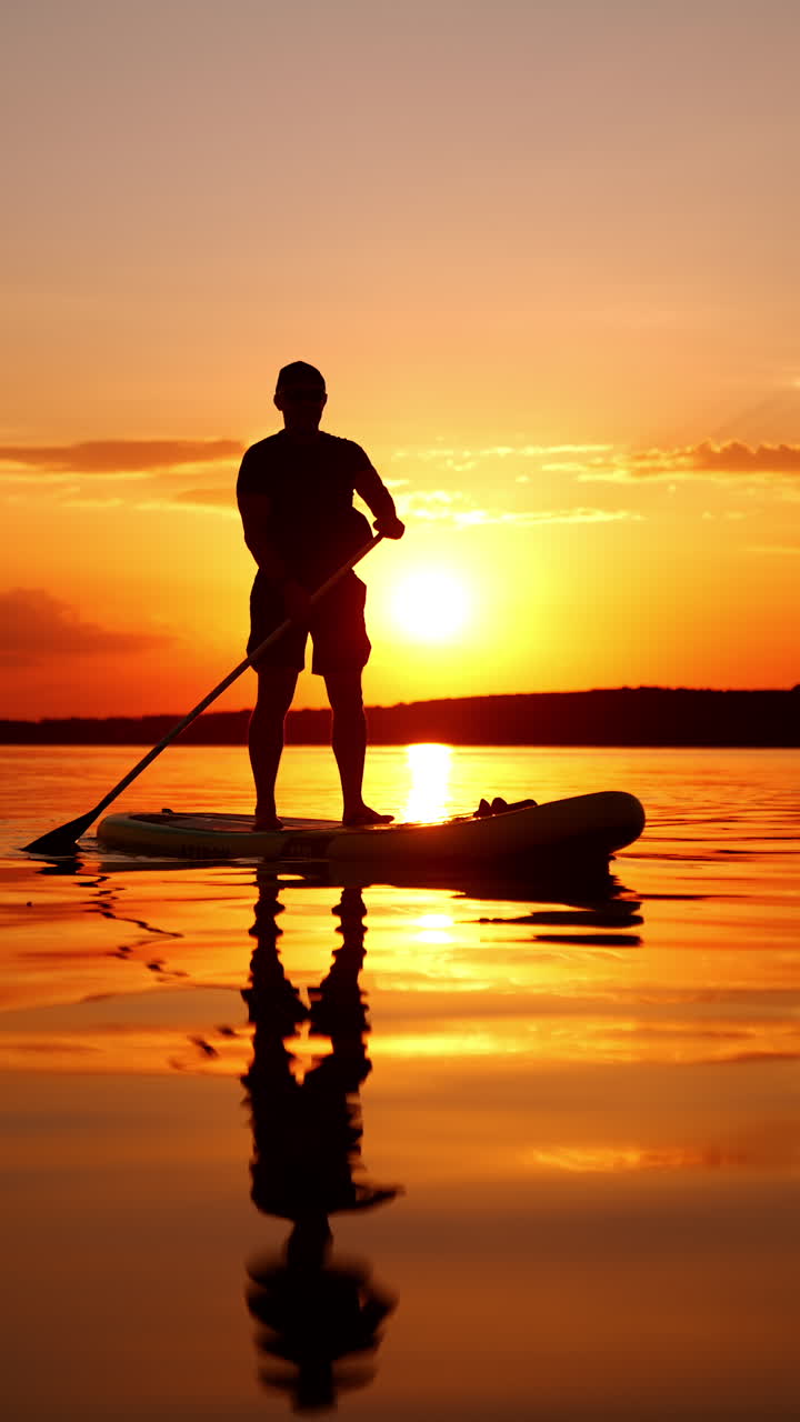 Male athlete standing on the sup board using an oar. Low angle view on the person paddle boarding on the river or lake at sunset. Vertical video.