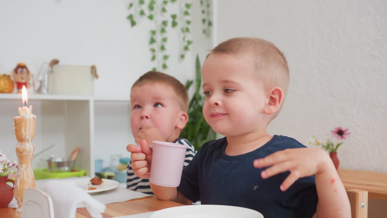 Boy with painted hand drinking from pink cup while sitting beside child eating food in striped shirt during kindergarten meal, surrounded by candlelight, plants, and soft daylight