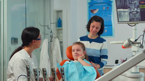 Niño-Usando-El-Dedo-Para-Señalar-El-Diente-Afectado-Mientras-El-Dentista-Habla-Con-La-Madre