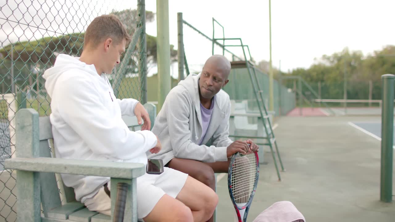 felices amigos varones diversos con raquetas de tenis hablando en el banco en la cancha de tenis en cámara lenta