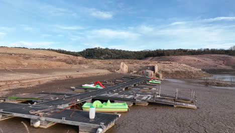 Vista-Aérea-Volando-A-Través-De-La-Plataforma-Del-Muelle-De-Canoa-De-Madera-Del-Pantano-De-Sau-En-Tierras-De-Sequía-Fangosas-En-La-Costa-De-Cataluña