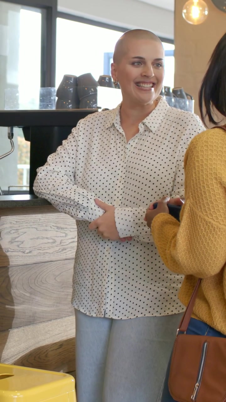 Video verticale: Donna con la testa rasata e camicia a pois che si sporge, ordinando un espresso al bancone del caffè, gesticolando.