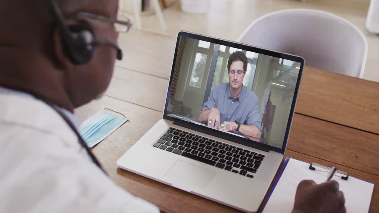 Afroamerikansk mandlig læge med headset på, der tager noter under et videoopkald på sin bærbare computer