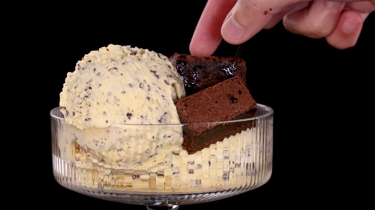 A hand places a chocolate brownie piece onto vanilla ice cream with chocolate chips in a glass dessert bowl, shot against a black background with bright, even lighting