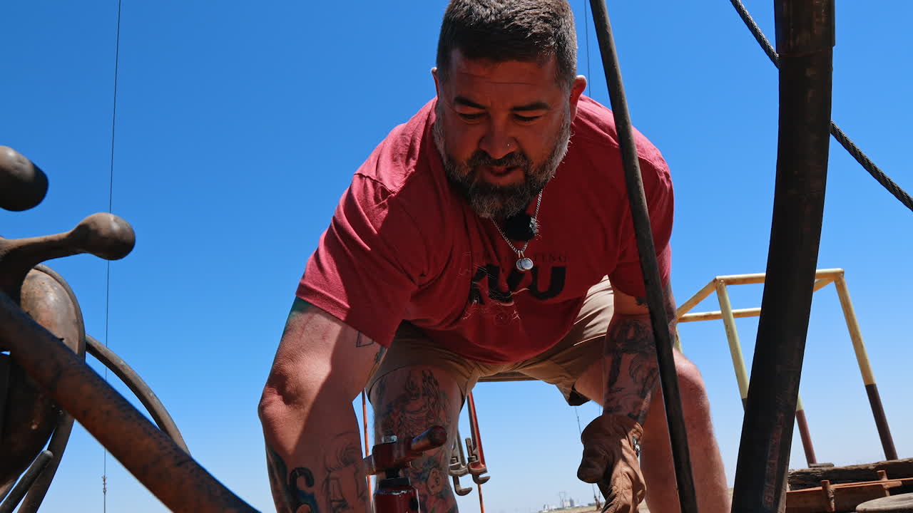 Bearded worker bent over the piece of pipe with a faucet. Low angle view at the man maintaining drilling equipment.